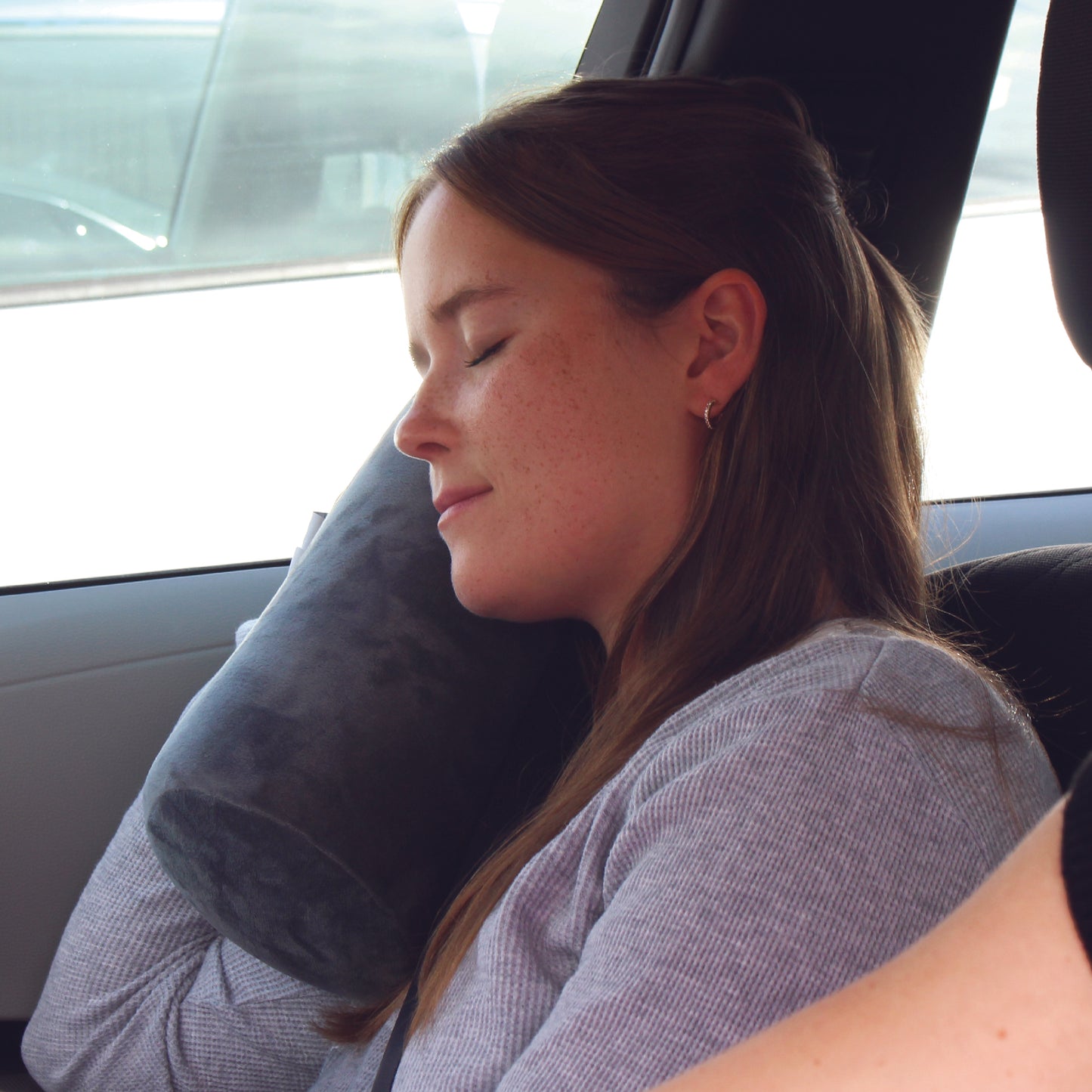 A woman traveling in the front seat of a vehicle holding the Plush Roll Travel Pillow near her neck while she relaxes and rests.