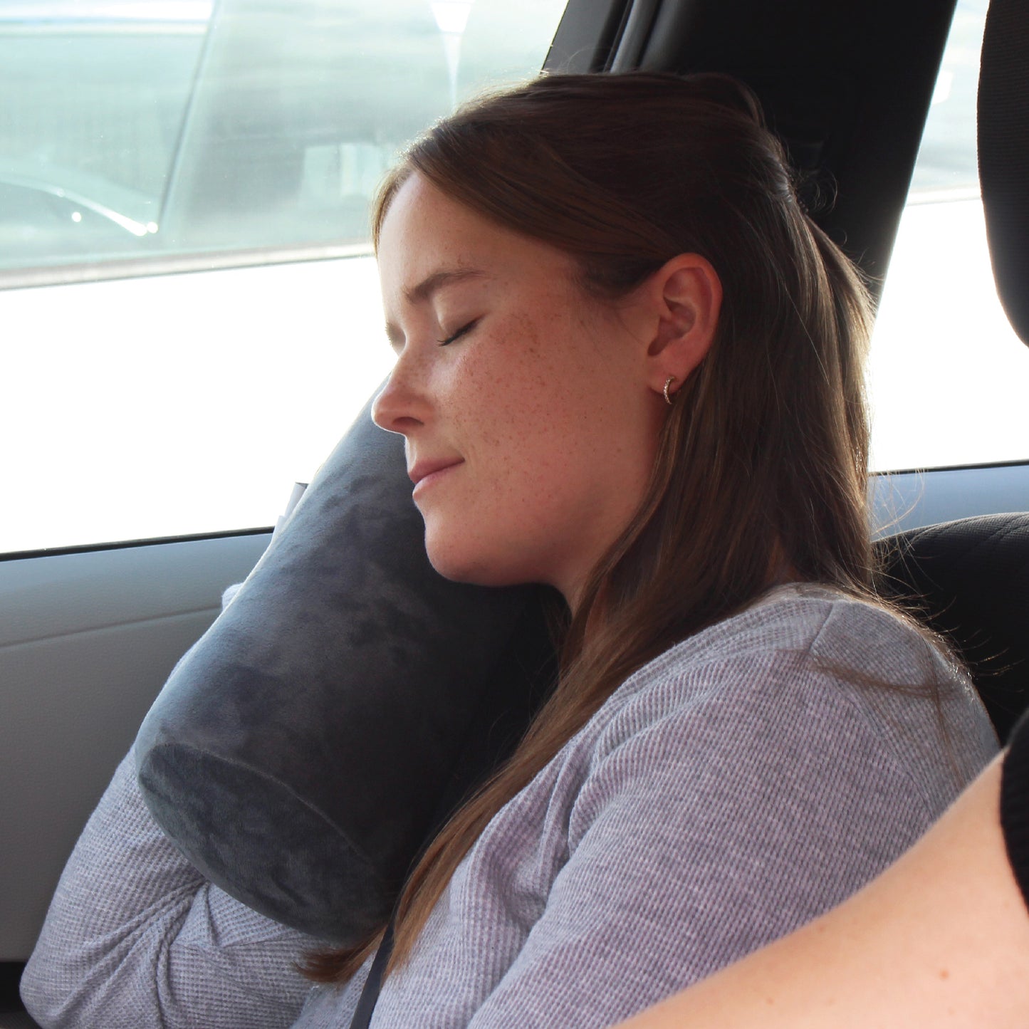 A woman traveling in the front seat of a vehicle holding the Plush Roll Travel Pillow near her neck while she relaxes and rests.