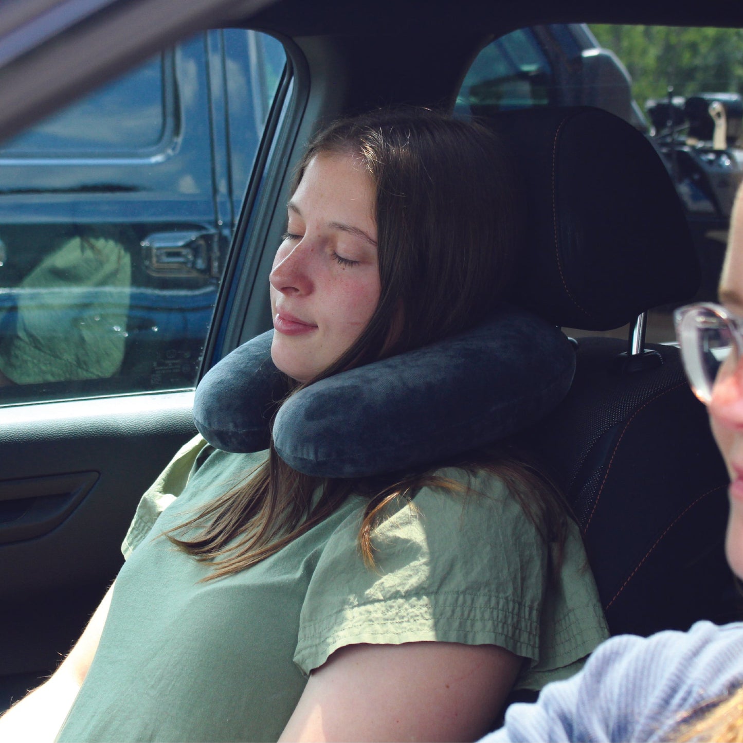 A woman traveling in the front seat of a vehicle wearing the Memory Foam Travel Neck Pillow around her neck while she relaxes and rests.