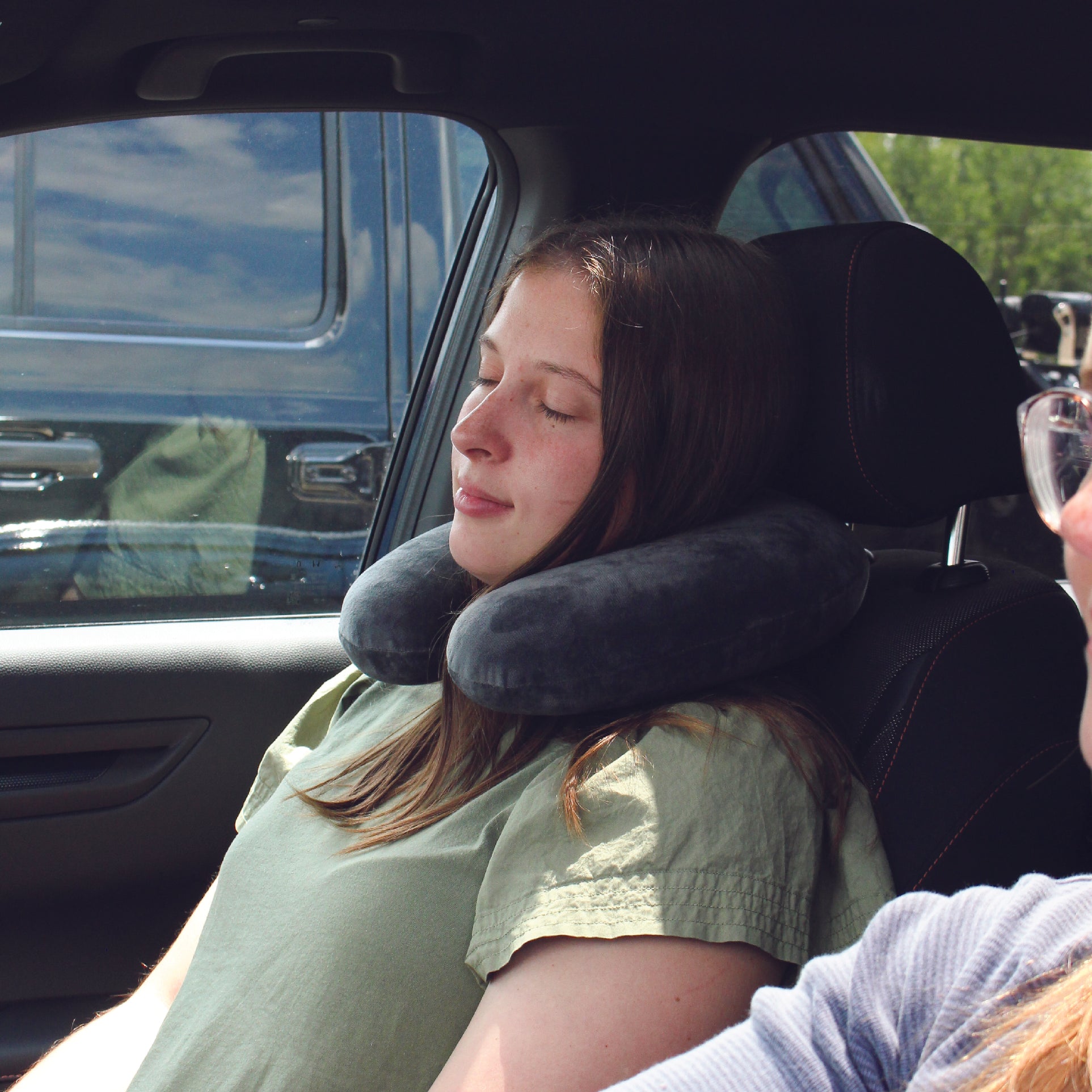 A woman traveling in the front seat of a vehicle wearing the Memory Foam Travel Neck Pillow around her neck while she relaxes and rests.