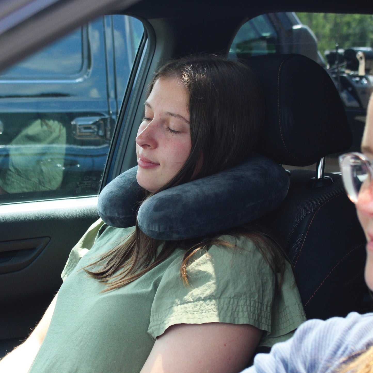 A woman traveling in the front seat of a vehicle wearing the Memory Foam Travel Neck Pillow around her neck while she relaxes and rests.