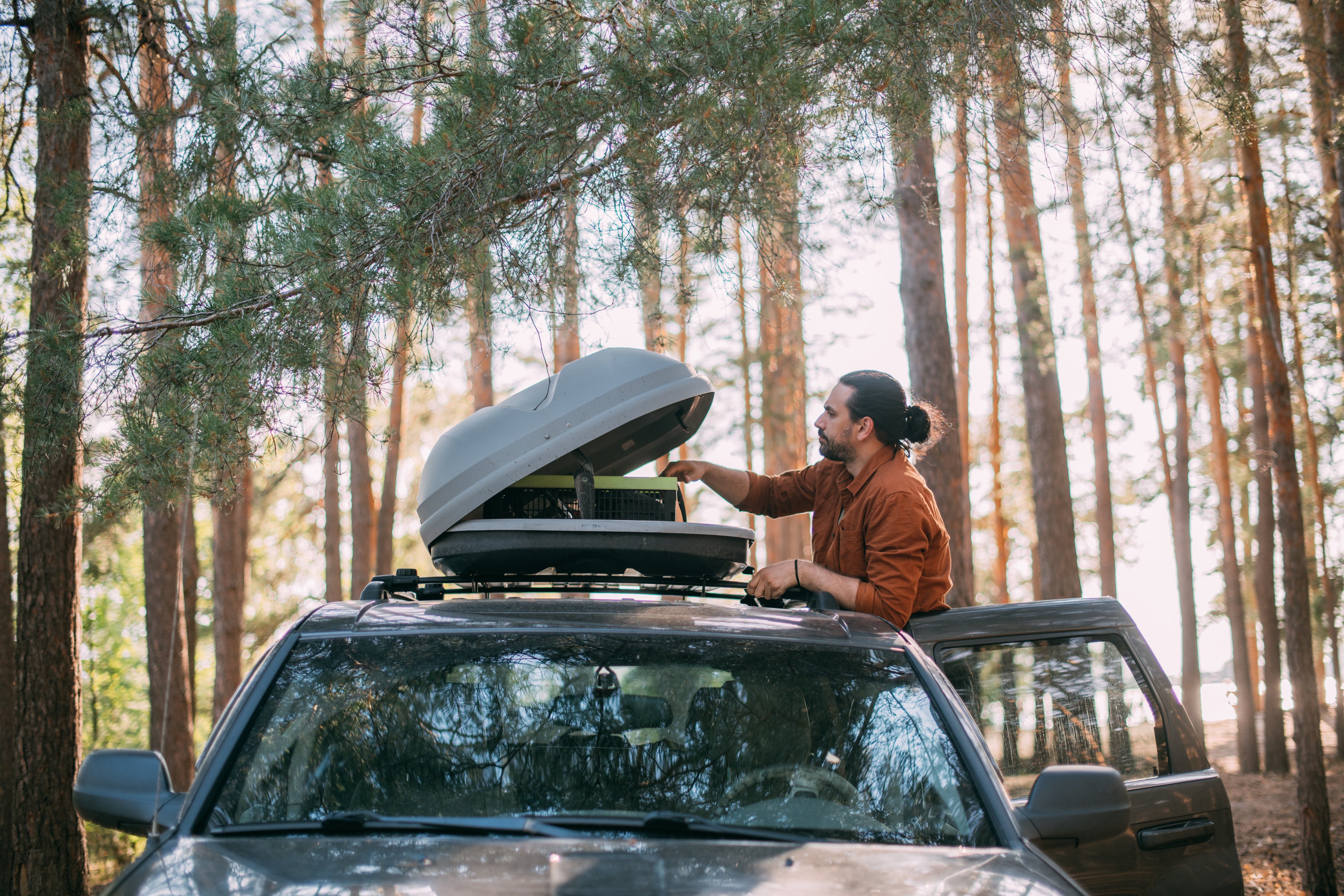 Traveler in a wooded area that has stepped outside their car to adjust a cooling unit inside a car-top storage container