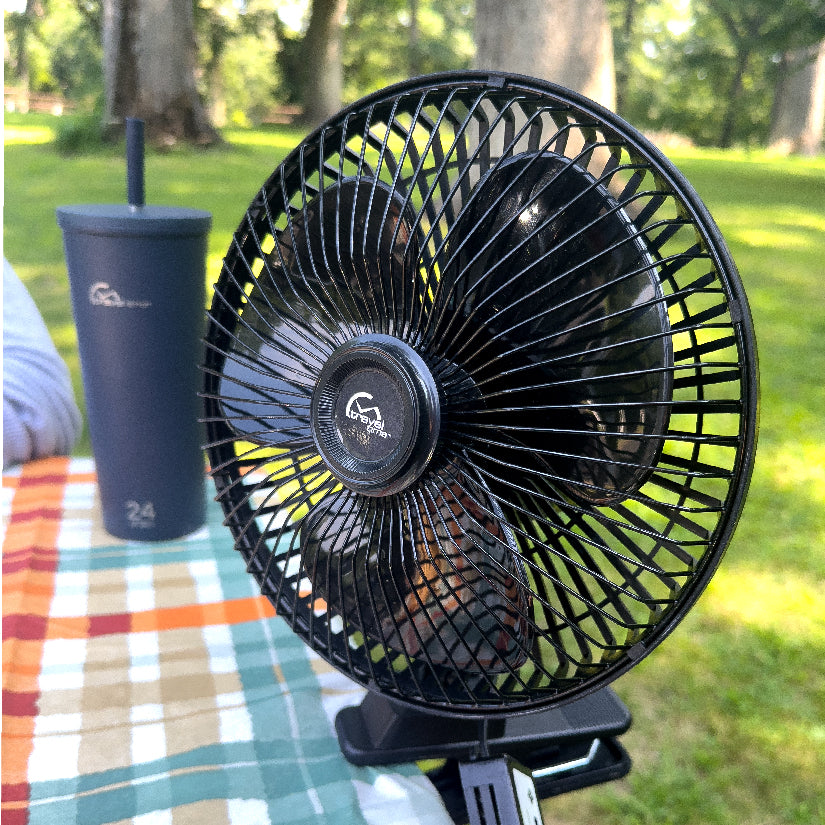 12V Oscillating Clip-On Fan clamped to a picnic table outside a campground, with a water bottle in the background