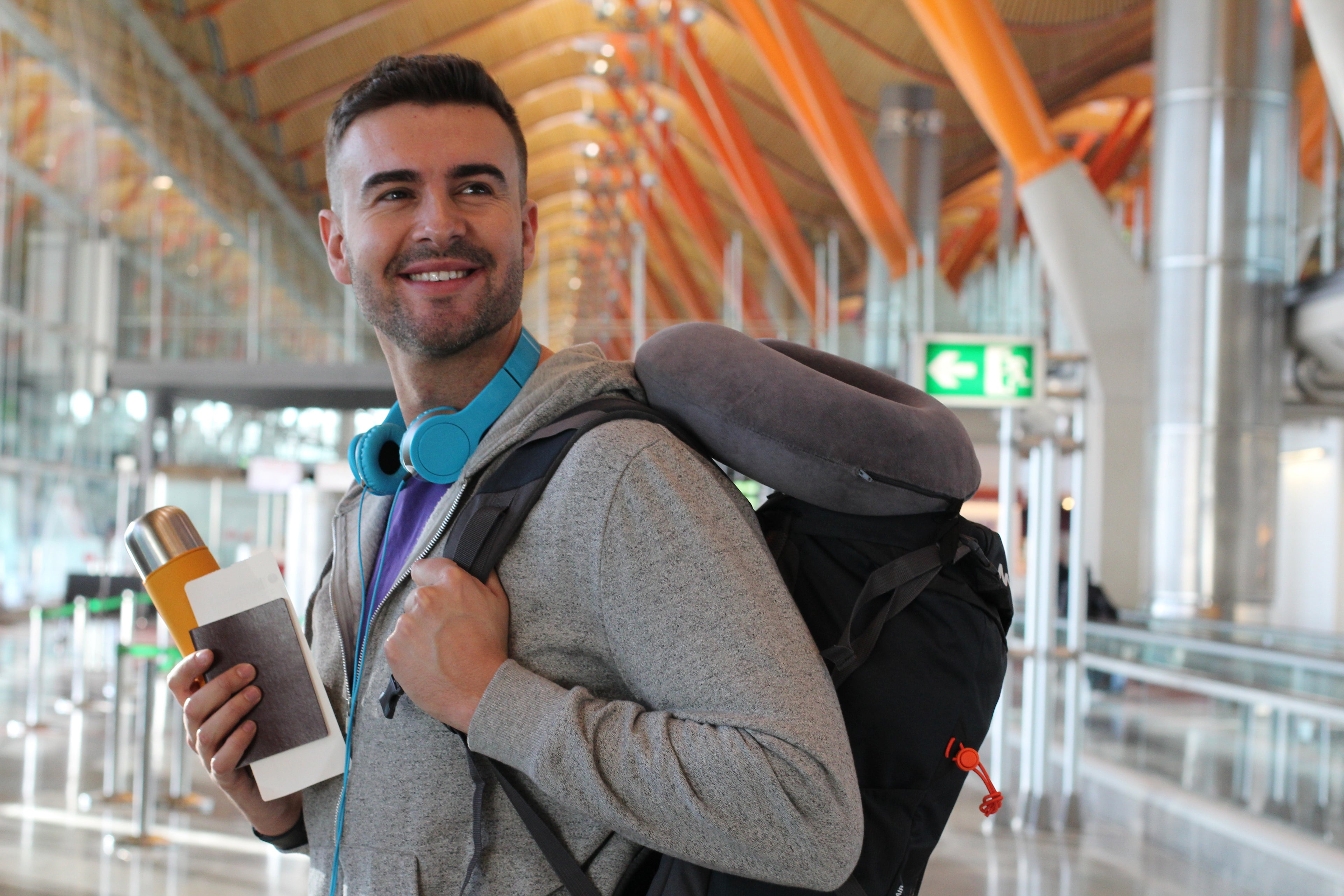 Traveller sporting a carry-on backpack with a Plush Travel Neck Pillow attached to it as they move throughout an airport. The traveller is also carrying his passport, water bottle, and boarding pass with a pair of bright blue headphones around his neck.