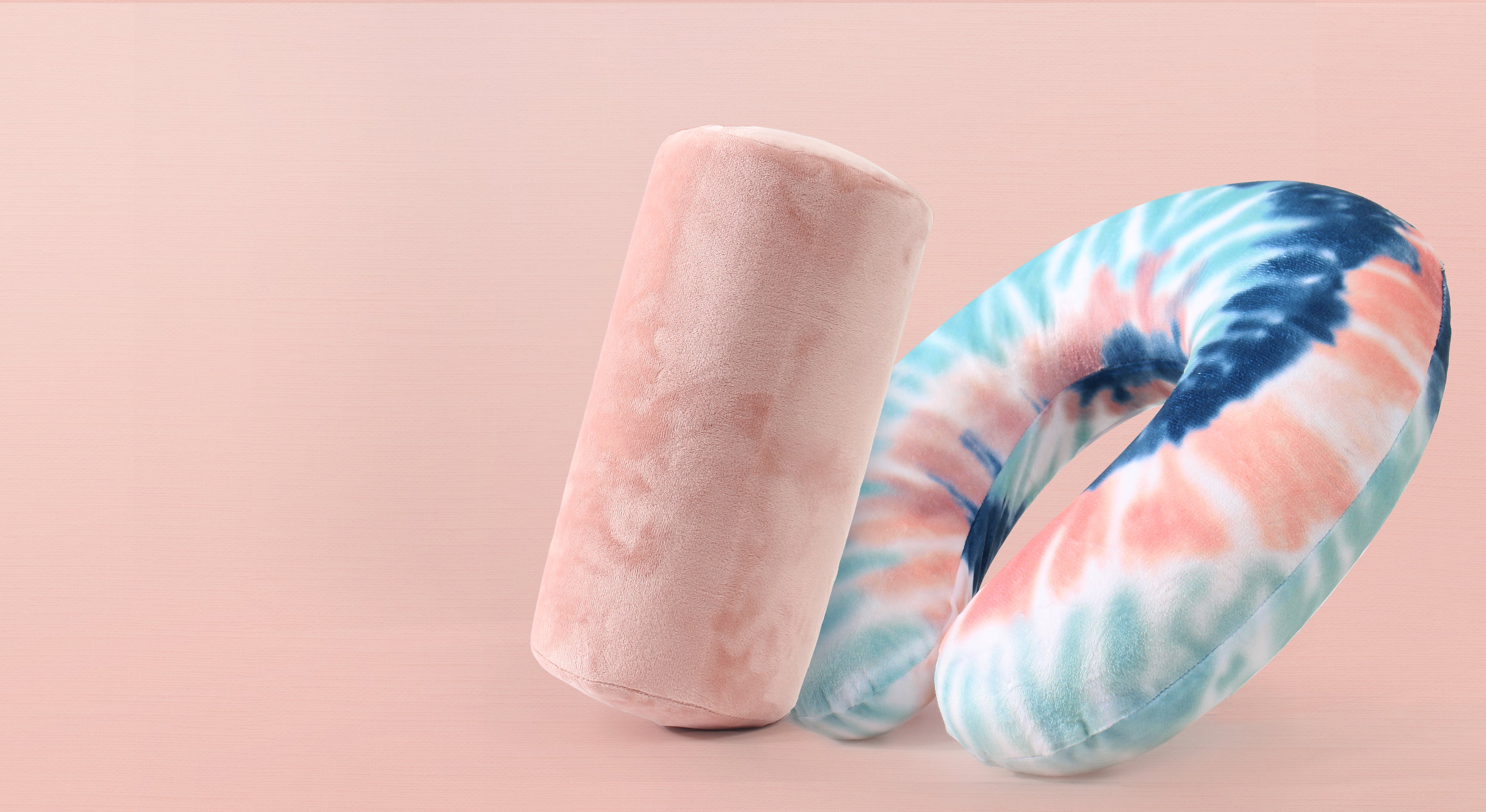 Memory foam roll travel pillow in pink and memory foam travel neck pillow in peach and gray tie dye, atop a light pink background
