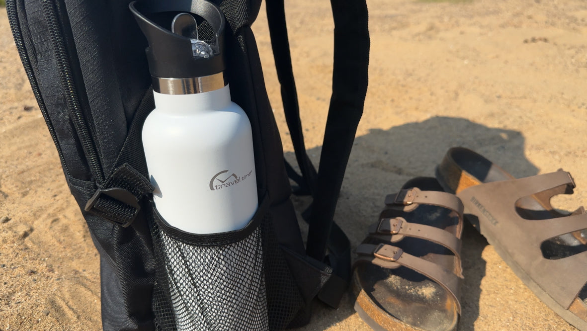 Stainless Steel Water Bottle with Flip Straw 20oz in White tucked into the side pocket of a backpack that is sitting in a sandy, beachy area