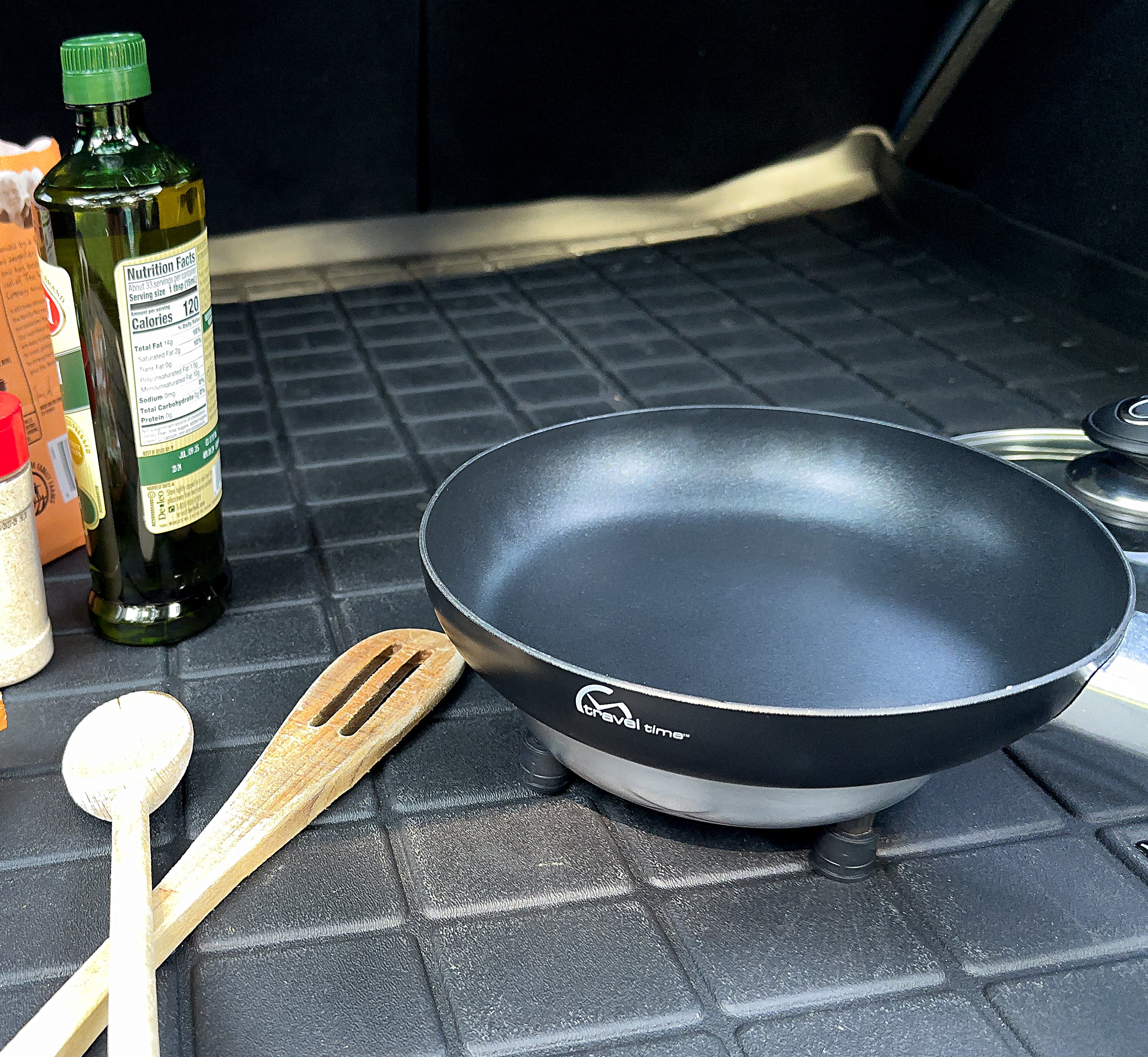 12 Volt Cooking Pan plugged into a 12 volt socket in the truck of a vehicle. Cooking utensils, olive oil, and seasoning are also visible, suggesting that a traveler is about to begin cooking a meal.